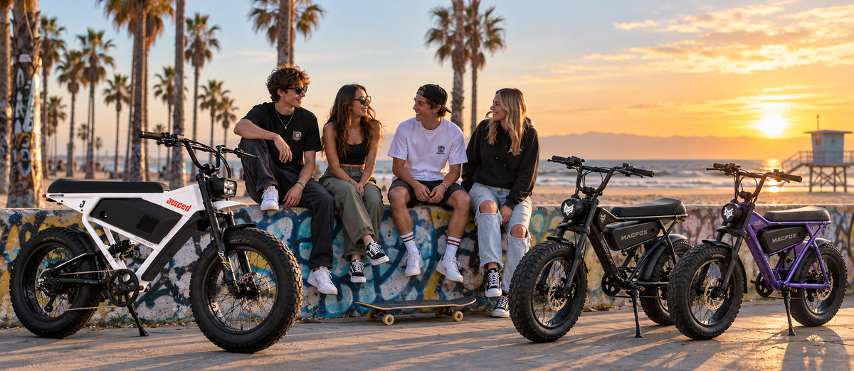 Three fat-tire electric bicycles are parked along a boardwalk at sunset, with four young people sitting on a colorful mural wall in the background. Palm trees and the ocean are visible behind them.